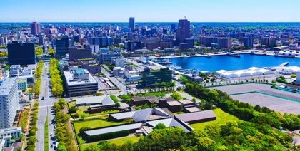 Cityscape of Chiba, Japan, featuring urban buildings, a waterfront harbor, and green spaces under a clear blue sky
