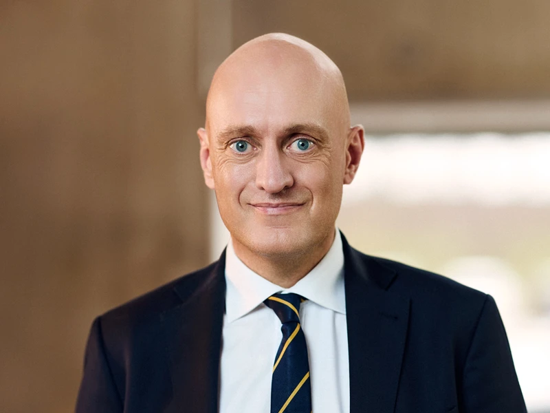 Mads Drejer, wearing a navy-blue suit and striped tie, stands confidently in front of a modern staircase