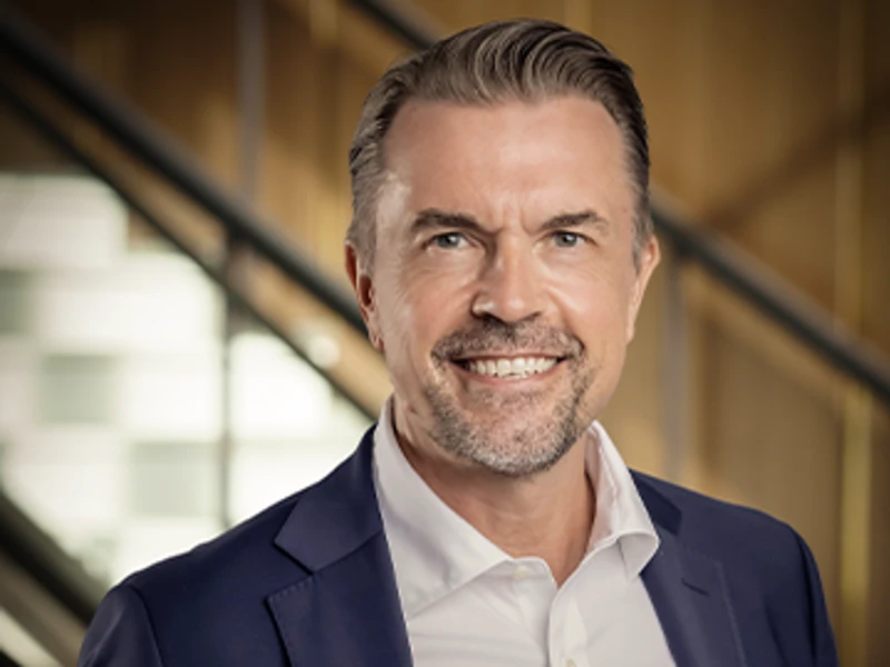 Rickard Ingvarsson in a professional portrait, wearing a navy-blue suit and white shirt, smiling confidently against a modern office background
