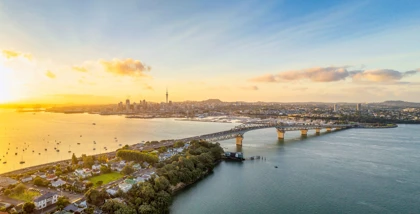 Aerial view of Auckland, New Zealand, featuring the Harbour Bridge, city skyline, and calm waters at sunset