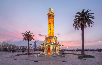The iconic Izmir Clock Tower stands illuminated in warm amber light at the center of Konak Square, Turkey, during a pastel-colored sunset. The ornate structure, built in 1901, features detailed Ottoman architecture with arches and a central clock face. Palm trees flank the tower, and people are seen strolling and gathering in the open plaza, highlighting the square’s cultural and historical significance