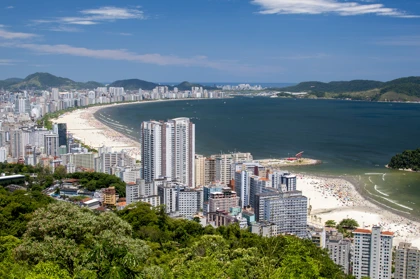 Aerial view of Santos, Brazil, showing a curved beach, tall residential buildings, and surrounding green hills under a clear blue sky