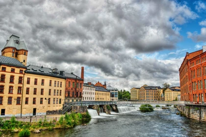 Scenic view of Norrköping, Sweden, featuring historic industrial buildings, a river, and cascading waterfalls under a cloudy sky