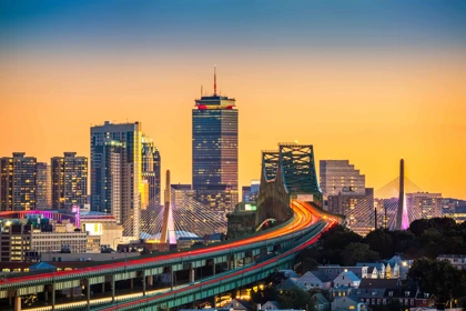 Boston skyline at sunset with the Tobin Bridge, Zakim Bridge, and Prudential Tower illuminated against an orange sky