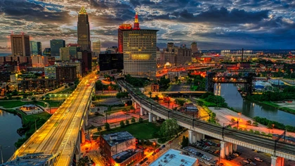 Nighttime aerial view of the Cleveland skyline with illuminated buildings, roads, and bridges reflecting on the Cuyahoga River