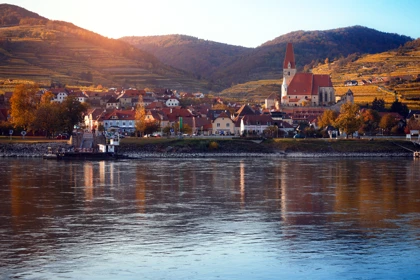 Scenic Austrian village with a red-roofed church, vineyards, and hills along the Danube River at sunset