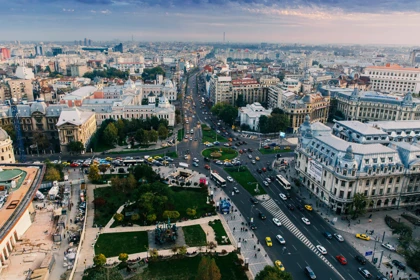 Aerial view of a bustling cityscape with a prominent intersection in Bucharest, heavy traffic, historic and modern buildings, green park with a statue, and partly cloudy sky