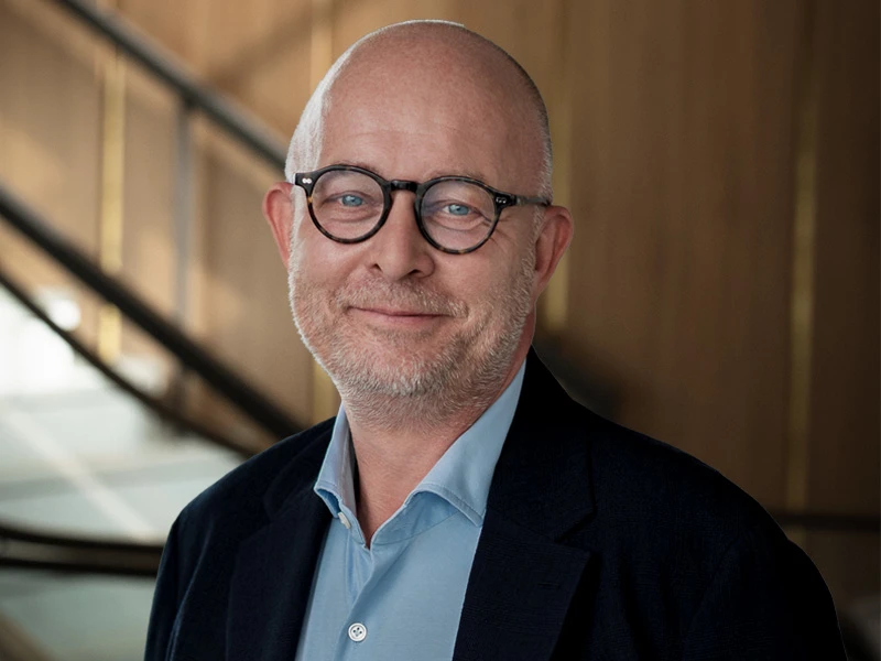 Lars Syberg in a professional portrait, wearing a dark suit, light blue shirt, and patterned tie, with glasses and a confident expression