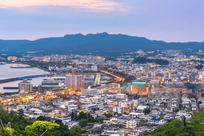 Evening cityscape of Okinawa, Japan, with coastal buildings, illuminated streets, and mountains in the background