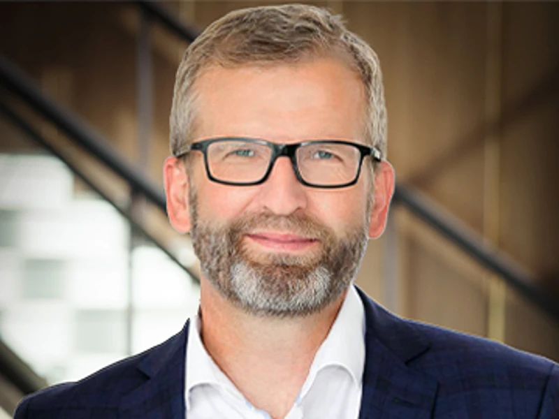 Morten Wagner, wearing glasses and a dark suit, smiles confidently in front of a modern staircase
