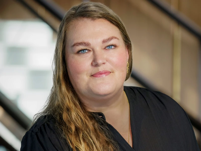 Birgitte Dam, wearing a black blouse, smiles confidently in front of a modern staircase