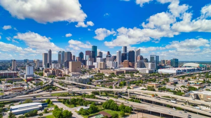 Downtown Houston skyline with modern skyscrapers, highways, and NRG Stadium under a bright blue sky with scattered clouds